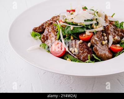 Salat mit warmem Rindfleisch, Tomaten, Rucola, Parmesan und Pinienkernen wird auf einem weißen Teller auf weißem Hintergrund serviert Stockfoto