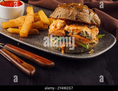 Lachsburger mit Blauschimmelkäse und Pommes frites wird auf einem grauen Teller mit Holzgriffen und Besteck serviert Stockfoto