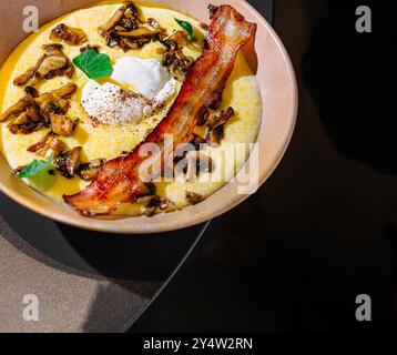 Köstliche Polenta mit gebratenen Pilzen, knusprigem Speck und pochiertem Ei, gewürzt mit frischen Kräutern, serviert auf einem Tisch Stockfoto