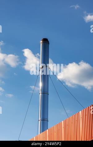 Ein sehr hoher gemauerter Kamin hebt sich von einem klaren blauen Himmel mit flauschigen weißen Wolken ab, was einen markanten Kontrast in der Stadtlandschaft schafft Stockfoto