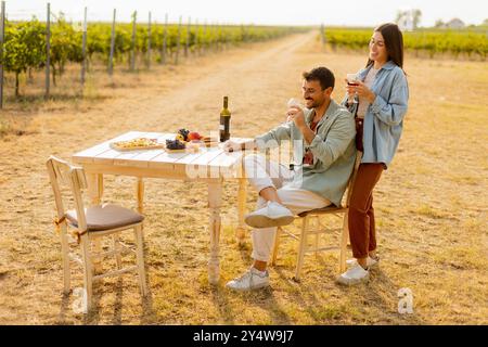 Ein paar entspannen sich an einem rustikalen Tisch in einem sonnigen Weinberg, genießen Wein und Gourmet-Snacks. Sie teilen Lachen und Freude, umgeben von üppigen Weinstöcken und dem Stockfoto