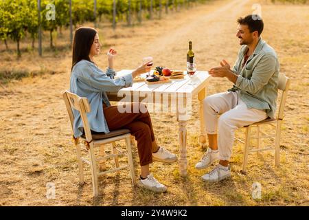 Ein paar entspannen sich an einem rustikalen Tisch in einem sonnigen Weinberg, genießen Wein und Gourmet-Snacks. Sie teilen Lachen und Freude, umgeben von üppigen Weinstöcken und dem Stockfoto