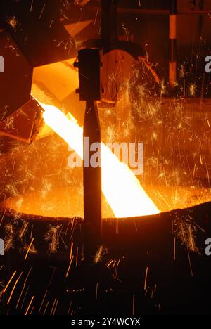 Dieses Bild zeigt den intensiven Moment, in dem geschmolzenes Metall aus einem Ofen in einen Behälter oder eine Form in einer Gießerei oder einem Stahlwerk gegossen wird. Der lebhafte oran Stockfoto