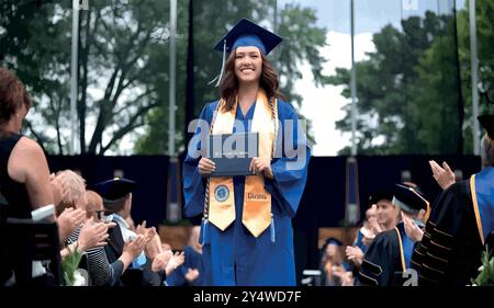 Eine stolze Hochschulabsolventin steht in ihrer Mütze und ihrem Kleid, hält ihr Diplom und lächelt, während sie nach vorne blickt. Sie markiert das Ende der harten Arbeit und einen Neuanfang. Stock Vektor