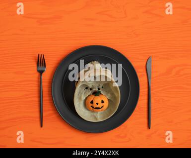 Halloween-Tisch mit Schädel auf schwarzem Geschirr und kleinem Kürbis auf orangem Hintergrund. Draufsicht, flach. Kopierbereich. Stockfoto