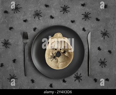 Halloween-Tisch mit schwarzen Geschirr und Schädel auf dunkelgrauem Hintergrund mit Spinnen. Blick von oben. Stockfoto