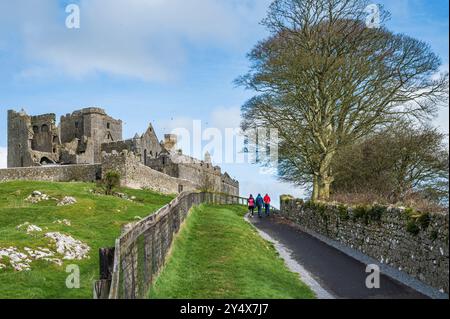 Blick auf den Felsen von Cashel, auf irisch; Carraig Phádraig, mit drei Wanderern, die entlang der Straße hinauf gehen. Cashel, Tipperary County, Irland. Stockfoto