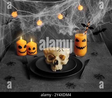 Halloween-Tisch mit Schädel in schwarzem Geschirr, brennenden Kerzen, Girlande auf dunkelgrauem Hintergrund mit Spinnweben. Stockfoto