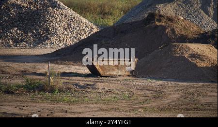 Ein verlassener rostiger Abfalleimer auf einer Baustelle, umgeben von Schotter- und Schotterhaufen, der Themen der industriellen Arbeit und Erdbewegungsarbeiten beleuchtet Stockfoto