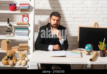 Porträt eines männlichen Lehrers aus der Vorschule oder Grundschule, der am Schreibtisch in der Klasse arbeitet. Reifer Mann sitzt entspannt in einem Klassenzimmer Blick auf die Stockfoto