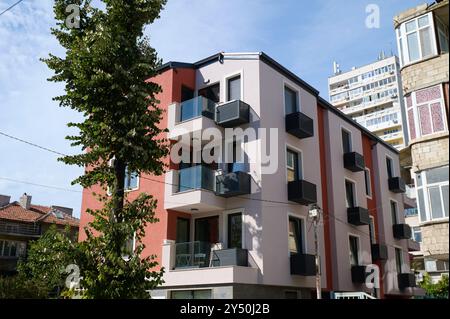 Ein neu erbautes Wohngebäude mit einer Mischung aus Farben und modernen Balkonen, vor einem Hintergrund von urbanem Grün bei hellem Tageslicht, EM Stockfoto