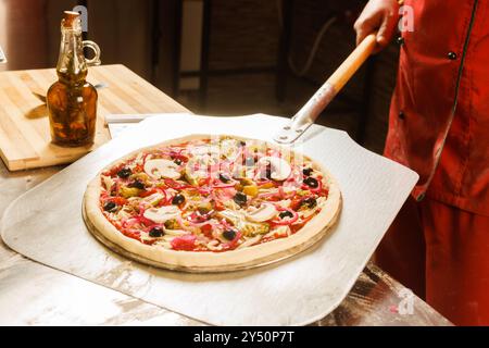 Ein Koch in roter Uniform bereitet sich darauf vor, eine Pizza mit Käse und Tomatenscheiben in einen Ofen zu stellen. Stockfoto