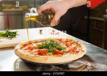 Ein handgegossenes Olivenöl auf eine frisch gebackene Pizza, garniert mit Rucola und Tomaten in der Küche. Stockfoto