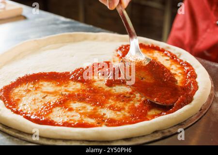 Eine Person, die Tomatensauce auf Pizzateig mit einem Kelle in einer Küche verteilt. Stockfoto