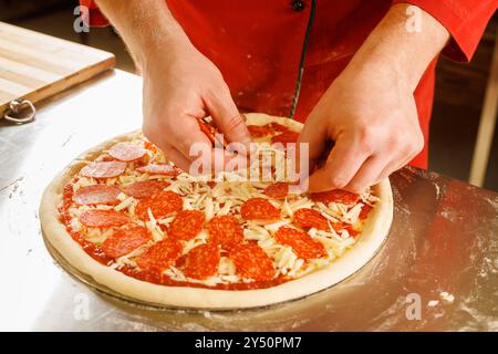 Eine Person in einem roten Hemd, die eine Peperoni-Pizza zubereitet, indem sie Peperoni-Scheiben auf Käse und Tomatensauce legt. Stockfoto