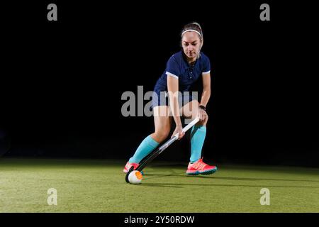 Weibliche Feldhockeyspielerin, die den Ball auf einem hellgrünen Rasen vor schwarzem Hintergrund dribbelt. Stockfoto