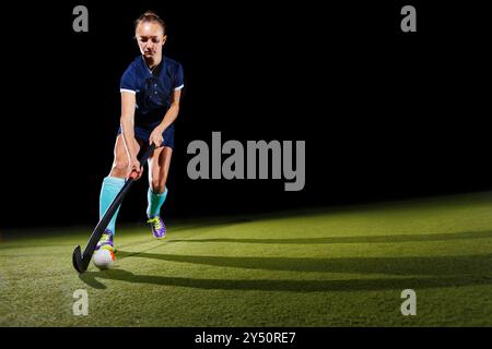 Eine entschlossene junge Frau, die den Ball geschickt mit dem Hockeyschläger anführt und sich auf ihr Spiel konzentriert. Isoliert auf schwarzem Hintergrund Stockfoto