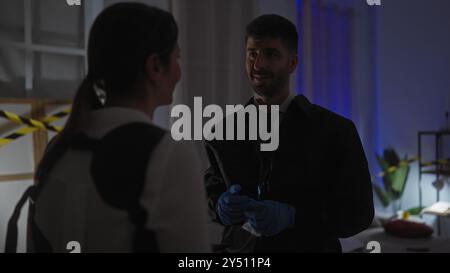 Ein Mann und eine Frau, beide Ermittler, untersuchen einen Tatort drinnen, mit Warnband und blauer Beleuchtung. Stockfoto