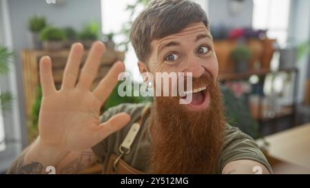 Fröhlicher bärtiger Mann, der in einem grünen Blumenladen winkt, mit Pflanzen im Hintergrund. Stockfoto