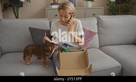 Eine junge blonde Frau, die in ihrem Wohnzimmer auf einem Sofa sitzt und mit ihrem Telefon spricht, während ein chihuahua-Hund an einer Paketbox auf der Couch schnüffelt. Stockfoto