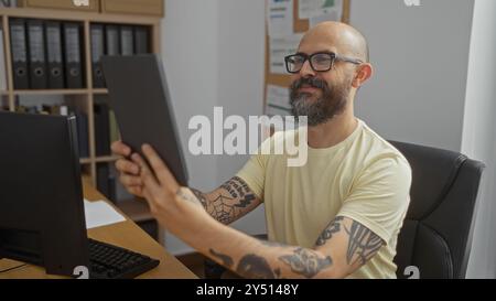 Hispanischer Mann mit Bart und Tätowierungen, der in einem Büro sitzt, schaut sich ein Tablet an einem Innenarbeitsplatz an. Stockfoto