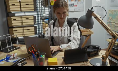 Eine zielgerichtete junge Frau untersucht einen Fall in einem überfüllten Polizeirevier, wobei sie ihren Laptop unter verschiedenen Beweisen benutzt. Stockfoto