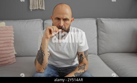 Hispanischer Mann sitzt in einem Wohnzimmer mit Tätowierungen auf den Armen, erscheint tief in Gedanken, während er ein weißes T-Shirt und Jeans trägt, in einem modernen Zuhause i Stockfoto