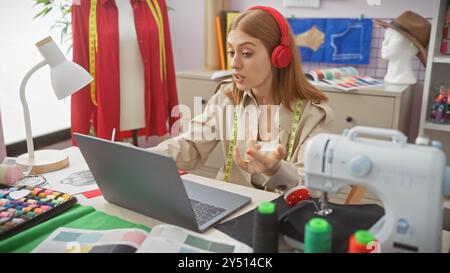 Junge Frau in Kopfhörer, die sich online berät, während sie in einem modernen Schneidergeschäft arbeitet. Stockfoto