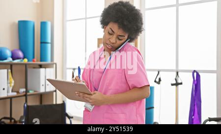 Fokussierte afroamerikanische Frau in rosa Peelings Multitasking mit Klemmbrett und Telefon in der Reha-Klinik. Stockfoto