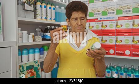 Eine ältere hispanische Frau in einem gelben Kleid untersucht Arzneimittelflaschen in einer Apotheke, umgeben von verschiedenen Produkten in den Regalen. Stockfoto