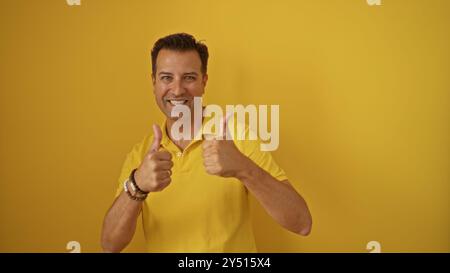 Gutaussehender, hispanischer Mann mittleren Alters, der lächelnd und mit zwei Daumen hoch vor einem gelben Hintergrund zeigt, was auf Positivität und Glück hindeutet Stockfoto