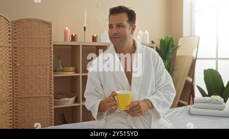 Hispanischer Mann, der sich in einem Spa-Wellnesscenter entspannen kann, während er einen gelben Becher hält, umgeben von Kerzen und beruhigender Einrichtung im Innenbereich Stockfoto