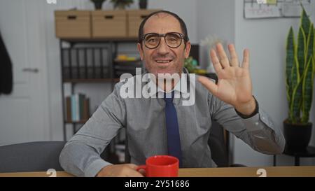 Älterer hispanischer Mann, der in einem organisierten Büro winkt, an einem Schreibtisch mit einer roten Kaffeetasse sitzt. Stockfoto