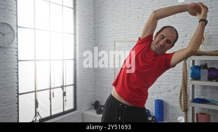 Hispanischer, kahler Mann mittleren Alters, der sich in einem Fitnessstudio mit Fitnessgeräten im Hintergrund an einem hellen, sonnigen Tag drinnen streckt. Stockfoto