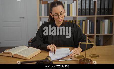 Attraktive hispanische brünette Richterin liest Dokumente in einem Gerichtssaal mit Hamelwaagen von Justiz und Gesetzbüchern Stockfoto