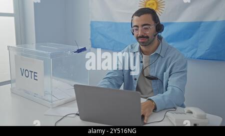 Junger Mann, der in einem Wahlbüro mit argentinischer Flagge im Hintergrund arbeitet, Kopfhörer trägt und an einer Wahlbox sitzt, die das veranschaulicht Stockfoto