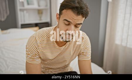 Junger hispanischer Mann mit Bart in gestreiftem Hemd, der nachdenklich auf dem Bett in einem Schlafzimmer sitzt. Stockfoto
