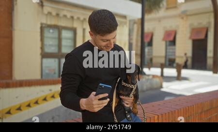 Ein junger hispanischer Mann mit seinem Dackelhund in einer städtischen Straße benutzt ein Smartphone. Stockfoto