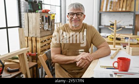 Lächelnder Mann mittleren Alters auf Schürze posiert in einer gut ausgestatteten Tischlerei mit Werkzeugen im Hintergrund. Stockfoto