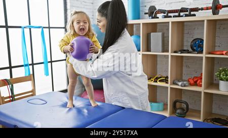 Ein Kinderarzt unterstützt ein junges Mädchen bei physiotherapeutischen Übungen mit einem Ball im Reha-Raum einer Klinik. Stockfoto