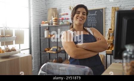 Eine junge Frau mit gekreuzten Armen steht selbstbewusst in einer Bäckerei, umgeben von Backwaren und einem Tafel-Menü Stockfoto