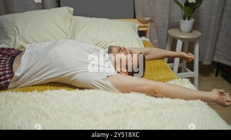 Junger hispanischer Mann mit Bart, der bequem auf einem Bett in einem gemütlichen Schlafzimmer liegt und seine Arme auf einer weichen Decke im Haus ausdehnt. Stockfoto