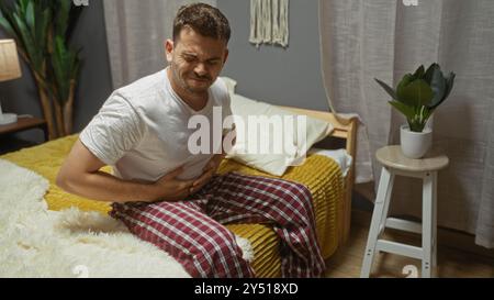 Ein junger hispanischer Mann im Pyjama sitzt auf einem Bett in seinem Schlafzimmer zu Hause, klemmt seinen Bauch vor Schmerzen, mit einer Pflanze und einem Beistelltisch, die in dem Platz sichtbar sind Stockfoto