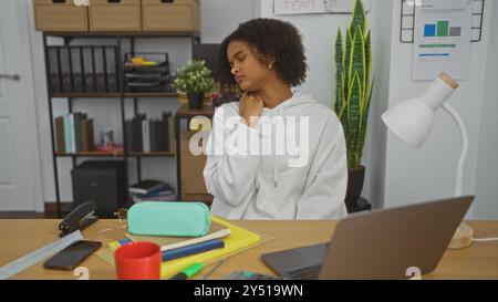 afroamerikanische Frau mit lockigem Haar in weißem Hoodie, die Nackenschmerzen hat, während sie an einem überdachten Schreibtisch mit Laptop, Zubehör und Muschel sitzt Stockfoto
