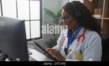 Eine schöne afroamerikanische Ärztin mit lockigen Haaren in einem Krankenhauszimmer, die ein Headset trägt und ein Tablet an ihrem Arbeitsplatz benutzt. Stockfoto