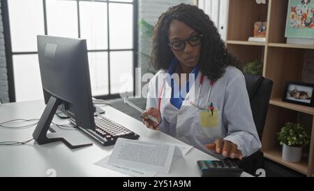 Eine junge afroamerikanerin in einem Klinikzimmer, als Arzt verkleidet, hält Geld, während sie an einem Computer und Rechner arbeitet. Stockfoto