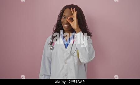 Eine schöne junge afroamerikanerin mit lockigem Haar macht eine gute Geste vor ihrem Auge, trägt einen weißen Arztmantel und steht gegen einen Stockfoto
