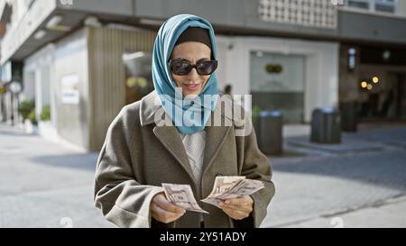 Eine reife Frau, die einen Hijab trägt, zählt saudische Riyals auf einer städtischen Straße im Freien. Stockfoto