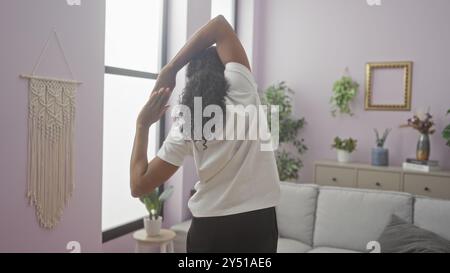 Eine schöne junge afroamerikanische Frau mit lockigen Haaren dehnt sich in einem gemütlichen Wohnzimmer aus, dekoriert mit Pflanzen und Makramee-Kunst, gebadet in natürlicher Sonne Stockfoto