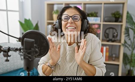 Hispanische Frau lächelt fröhlich während der Aufnahme in einem modernen Radiostudio mit professionellem Mikrofon und Kopfhörern. Stockfoto